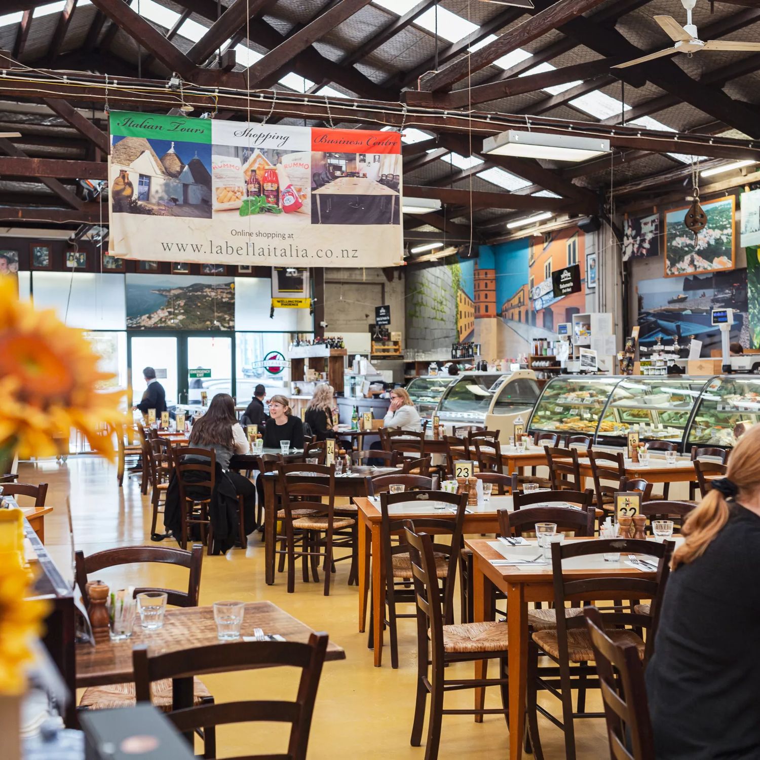 Interior of Bella Italia in Petone, showing a spacious dining hall with wooden tables and chairs, an open deli-style counter, and sunflowers in the foreground.