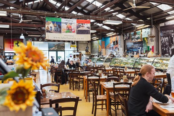 Interior of Bella Italia in Petone, showing a spacious dining hall with wooden tables and chairs, an open deli-style counter, and sunflowers in the foreground.