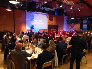 Guests fill a room inside Prefab Hall for a conference, as a speaker stands on a podium. the lighting in the room is dim and blue.