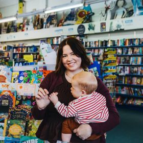 Hannah Carr holds a baby while browsing in a bookstore, with colourful children’s books and shelves filled with books in the background.
