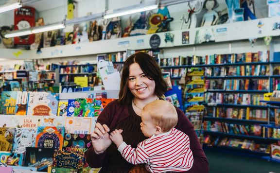 Hannah Carr holds a baby while browsing in a bookstore, with colourful children’s books and shelves filled with books in the background.