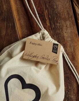 A light beige canvas drawstring bag with an illustrated black heart printed on it laying on a wooden surface with a brown rectangular cardstock placed on it that reads "Enjoy this: Wellington Goodies Bag". 