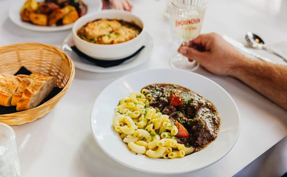 Beef bourguignon served on a white tablecloth alongside a basket of bread, a bowl of French onion soup, and duck confit.
