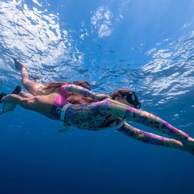Two people dive under the water wearing colourful wet suits. They wear goggles and flipers.