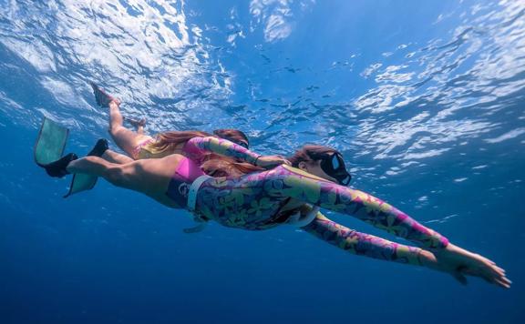 Two people dive under the water wearing colourful wet suits. They wear goggles and flipers.