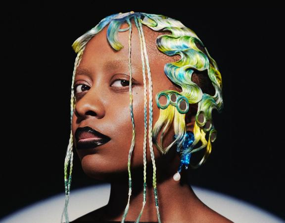 2023 Wellington Jazz Festival performer Cécile McLorin Salvant looking directly at the camera. She has bright green and blue hair with exotic styling and thing plaits hanging down her face.