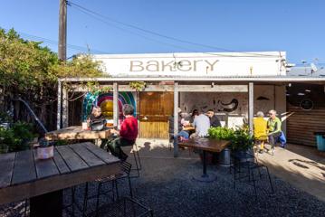 An exterior seating area at  Olde Beach Bakery in Waikanae. Three groups of people sit at tables enjoying coffee and pastries.