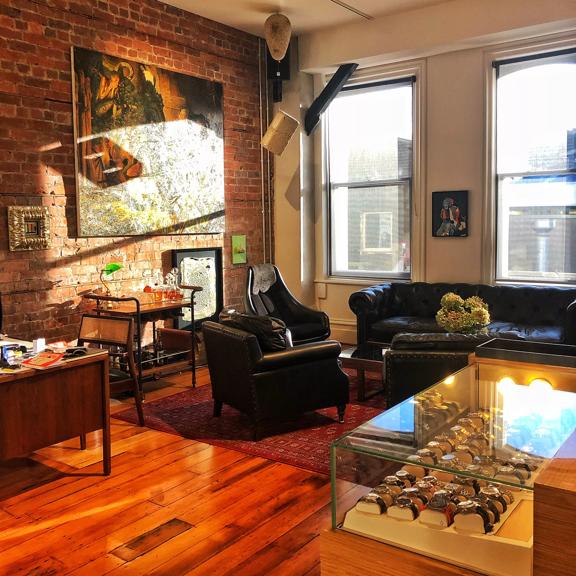 Interior shot of Five:45. Large sash windows are in the back ground letting and sunlight is streaming through. A brick wall is on the left had side hung with art work. Couches, a coffee table, a desk, and drinks trolley can be seen in the foreground.