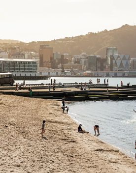 Oriental Bay Beach in Wellington. There are people on the beach, in the water and on the pier and the city's downtown is visible in the background.