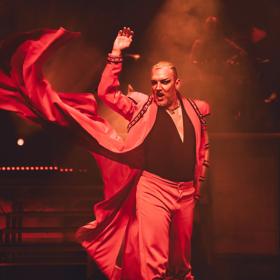 An actor performing in Dracula's: Sanctuary, an Australian cabaret show. They are centre-stage, singing and dramatically whipping their red cape.