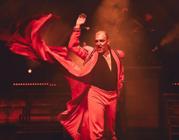 An actor performing in Dracula's: Sanctuary, an Australian cabaret show. They are centre-stage, singing and dramatically whipping their red cape.