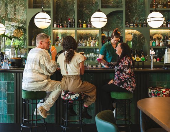 Interior of Hummingbird restaurant in Wellington, with guests seated at a green-tiled bar while a staff member prepares drinks behind the counter, shelves of bottles and glassware illuminated by warm, ambient lighting.