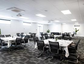 The Bond conference room at NZCIS. The tables are set for an event with white tablecloths and glasses. There are white walls and ceiling and grey carpet.
