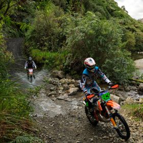Two riders on motorbikes passing through a stream on the Karapoti Challenge course