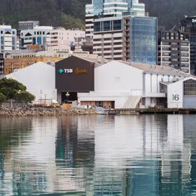 Looking across the waterfront at the TSB arena and Shed 6, 3 large sheds painted white and 1 painted black. The CBD buildings can be seen behind.