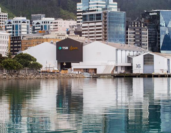 Looking across the waterfront at the TSB arena and Shed 6, 3 large sheds painted white and 1 painted black. The CBD buildings can be seen behind.