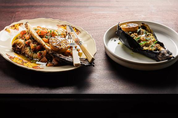 2 plates of food on a wooden table at Cinderella, one a stuffed eggplant served with lemon and the other a roast dish.