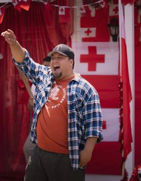 A still from the film Red White and Brass with a person wearing a blue plaid shirt, orange tee shirt, black pants and a black baseball cap stands raising his arm into the air with an elated expression.