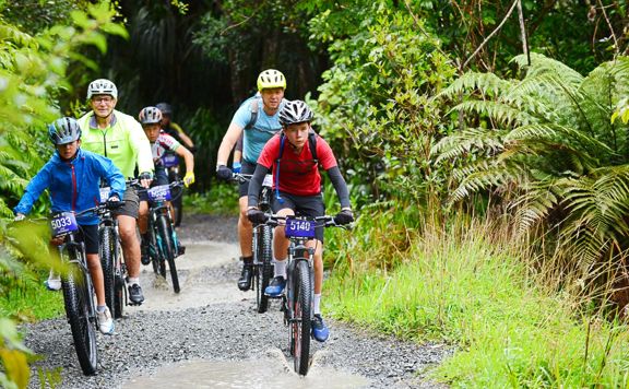 A group of mountain bikers ride side-by-side along a gravel path with puddles. They are surrounded by lush green bush.
