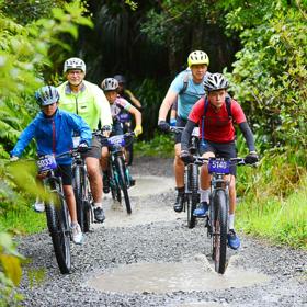 A group of mountain bikers ride side-by-side along a gravel path with puddles. They are surrounded by lush green bush.