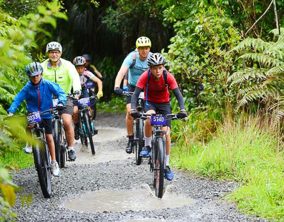 A group of mountain bikers ride side-by-side along a gravel path with puddles. They are surrounded by lush green bush.