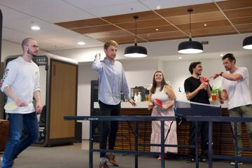 Two men play ping pong in a modern office kitchen with three colleagues standing in the background.