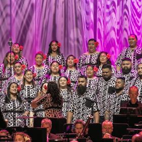 A Pacific community choir performs with live accompaniment in front of a curtain with pink decorative lights. 