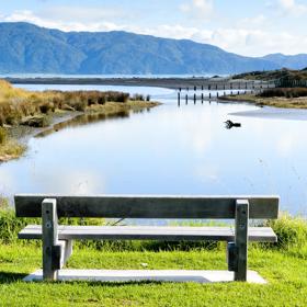 A grey wooden bench faces a pond in Waikanae Estuary Scientific Reserve in Paraparaumu, New Zealand. There is tall grass, a piece of driftwood and mountains visible in the background. 