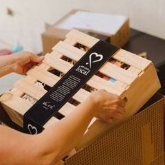 A person who is out of frame places a wooden box with a black label wrapped around it into a larger cardboard box.