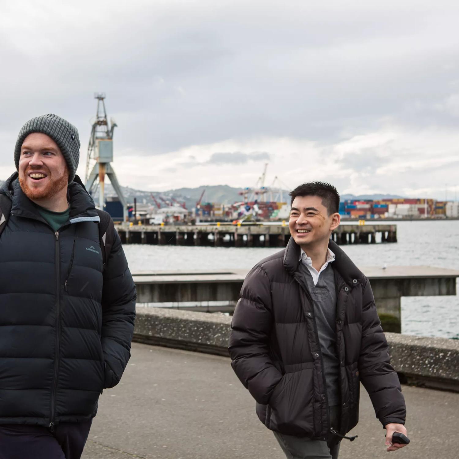 Two smiling people wearing dark-coloured puffer jackets walk along the habour front in Wellington on an overcast day. 