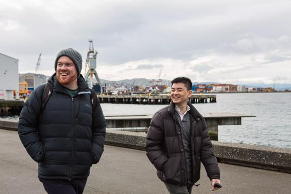 Two smiling people wearing dark-coloured puffer jackets walk along the habour front in Wellington on an overcast day. 