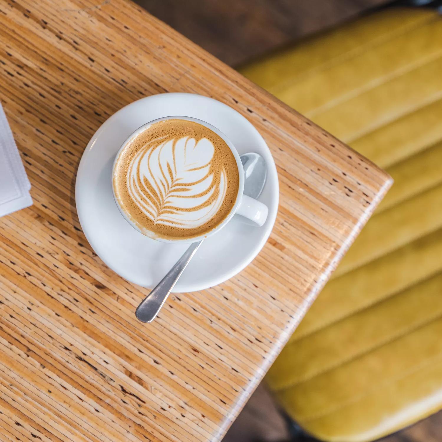 A bird's eye view of a foamy cappuccino in a white coffee cup on a white saucer sitting on a light beige table top at Swimsuit Cafe located on Dixon Street in Wellington.