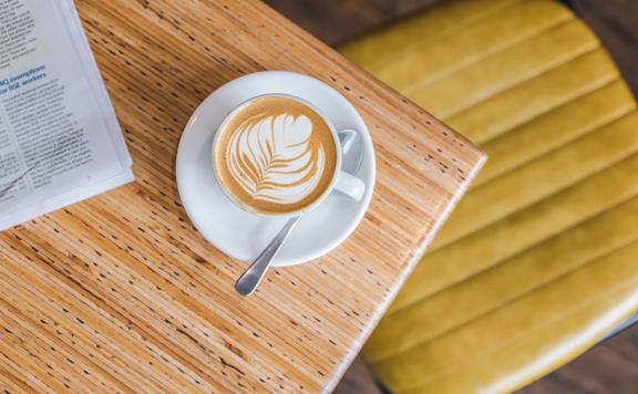 A bird's eye view of a foamy cappuccino in a white coffee cup on a white saucer sitting on a light beige table top at Swimsuit Cafe located on Dixon Street in Wellington.