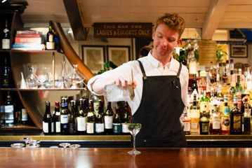A bartender wearing a black apron and white collared shirt pours a cocktail into a glass.