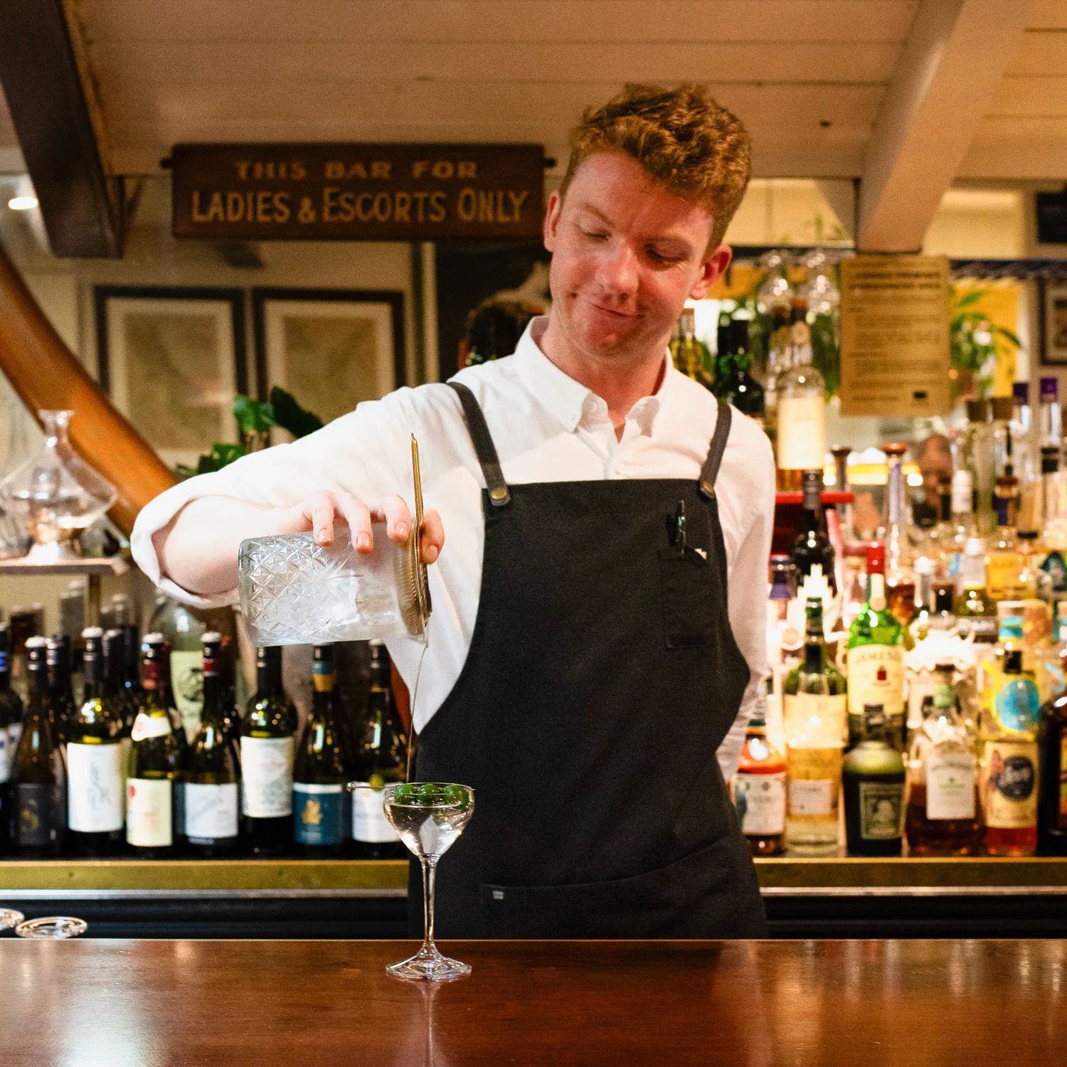 A bartender wearing a black apron and white collared shirt pours a cocktail into a glass.