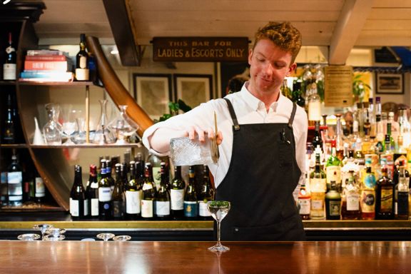 A bartender wearing a black apron and white collared shirt pours a cocktail into a glass.
