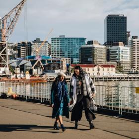 Two people walk along Wellington's waterfront on a sunny winter day. The city centre is visible in the background.