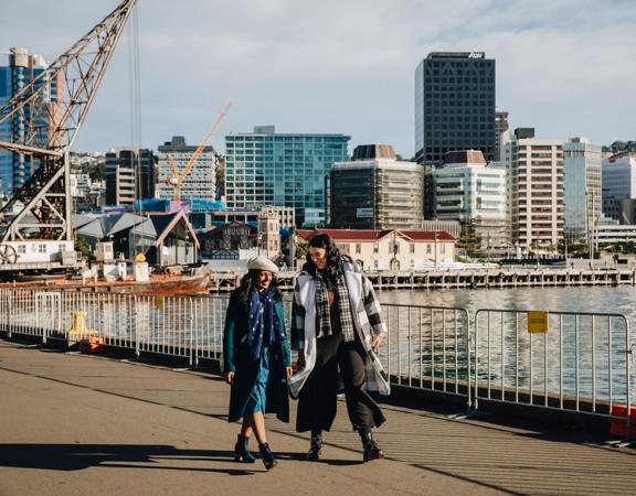 Two people walk along Wellington's waterfront on a sunny winter day. The city centre is visible in the background.