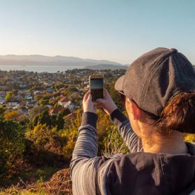 A person stands, taking a picture with their phone of a scenic view over the city of Wellington.