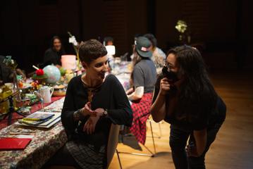 Two people conversing while another four people sit at a table in the background in a dark room.