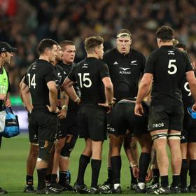 The All Blacks rugby team huddles on the field with fans blurred out in the background.