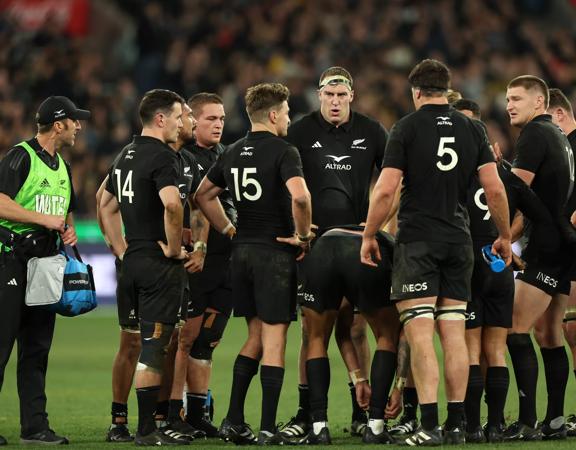 The All Blacks rugby team huddles on the field with fans blurred out in the background.