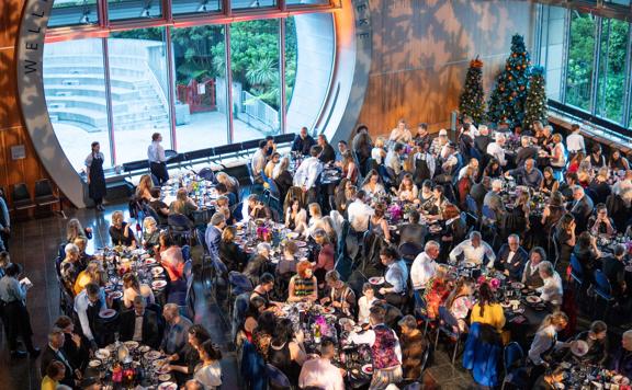 A large Christmas themed event in the foyer of Te Papa Tongarewa, people are sat at tables enjoying food and a large window shines light on them.