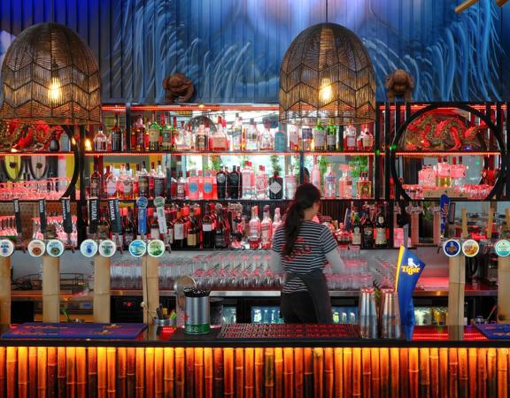 A bartender faces the wall while standing behind a colourful, tiki-style bar.