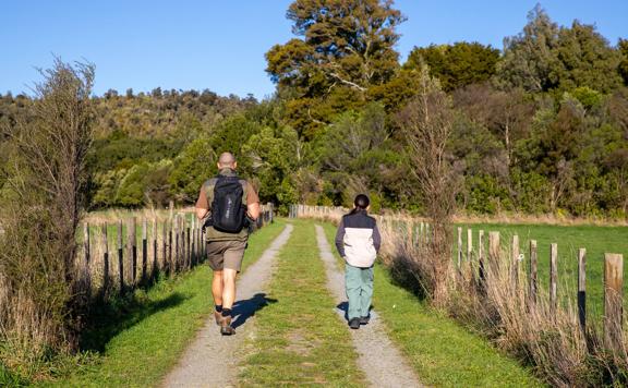Two people walking along a grassy and gravel trail towards the forest at Fensham Reserve in Wairarapa, with wooden farm fences lining the track.