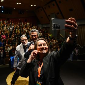 Three people take a selfie in front of a packed audience.