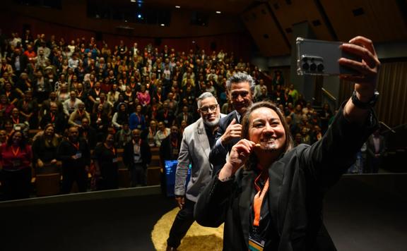 Three people take a selfie in front of a packed audience.