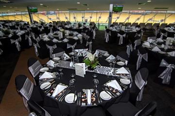 Inside a corporate box at Hnry Stadium Function Centre. Many tables are set with silverware and rugby playbook. The large glass windows at the edge of the room overlook the rugby field.