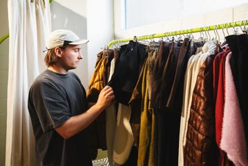 Person browsing through a clothing rack at the Coffee Outdoors Wellington store, holding a pair of black pants among various garments including jackets.