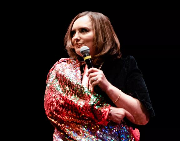 Deborah Frances-White, host of the award winning podcast 'The Guilty Feminist', on stage holding a microphone wearing  a sparkly shall.